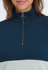 Maglia navy con zip e colletto, dotata di una zip argento e un pannello inferiore grigio. Tessuto a maglia liscia con una breve zip al collo per una chiusura regolabile.
