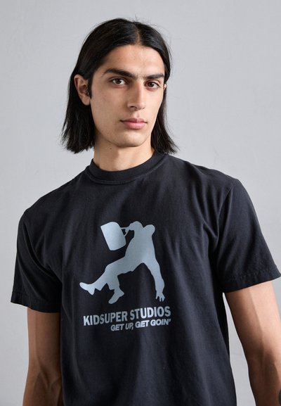 Sort bomulds T-shirt med et sølv grafisk motiv af en person, der holder en beholder, med teksten "KIDSUPER STUDIOS" og "GET UP, GET GOIN’" nedenfor.