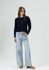 Marineblauwe gebreide cardigan met ronde hals en knoopsluiting, gecombineerd met lichtblauwe wijde jeans. Model draagt tan instappers.