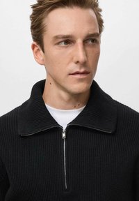 Maglione nero a costine con colletto con zip e una maglietta bianca sotto. La zip ha un tirante argentato e il tessuto ha una finitura testurizzata.
