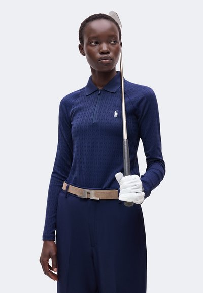 Femme vêtue d'une tenue de golf bleu marine et d'un gant blanc tenant un club de golf vertical à côté de son visage, regardant sur le côté sur un fond uni.