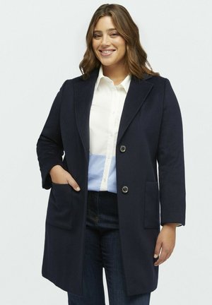 Cappotto di lana navy con un design su misura, caratterizzato da un colletto a revers, due tasche frontali e due bottoni. Indossato sopra una camicia bianca e jeans scuri.