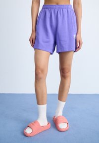 Lila bomullsshorts med elastisk midja, sidofickor och vit logotyp, i kombination med rosa slidesandaler och vita mässingsockor.