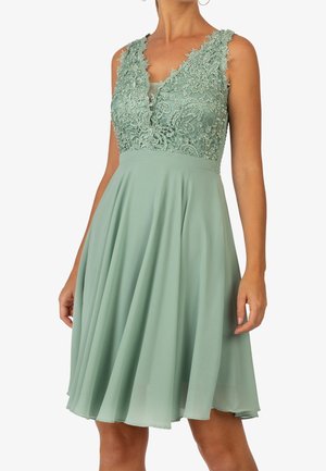 Ærmeløs salviegrøn kjole med blonder blomster bodice, V-udskæring og flydende chiffon nederdel, der går til over knæene, båret af en stående person.