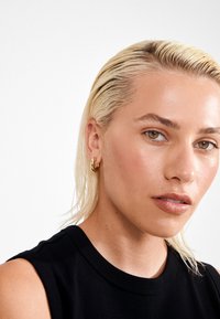 Ung person med vådt blondt hår, guld hoops øreringe, iført en sort ærmeløs top, som ser lidt til side mod en hvid baggrund.