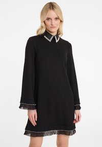 Robe noire à manches longues, col orné de perles, et poignets et ourlet texturés et plissés. Matière lisse avec une silhouette simple.