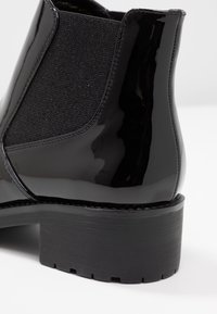 Černé kotníkové boty s lesklým svrškem z lakované kůže, elastickým bočním panelem, texturovaným černým podpatkem a silnou gumovou podrážkou s vzorkem.