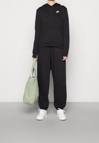 Svart hoodie med framficka och logotyp, kombinerat med lösa svarta byxor. Vita sneakers och en grön tote-väska fulländar outfiten.