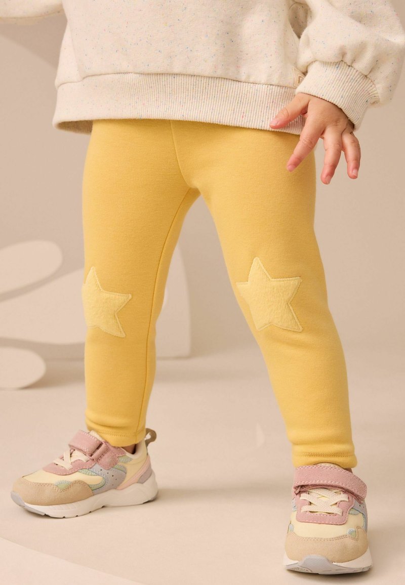 Enfant portant un legging jaune avec des patchs en forme d’étoile aux genoux et des baskets multicolores avec des brides roses, debout sur un fond neutre.