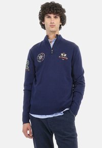 Navy blue zip-up sweater in knit fabric, featuring embroidered logos and patterns on the chest and sleeves. Long sleeves, ribbed cuffs.