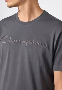 Mężczyzna w ciemnoszarej koszulce polo z krótkim rękawem, z haftowanym logo "Champion" na klatce piersiowej, zwrócony w lewo.