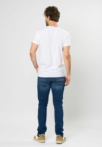Fehér, rövid ujjú póló kék, slim-fit farmernadrággal és bézs tornacipővel viselve. A háttér semleges szürke, amely kiemeli az outfittet.