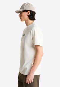 Joven con gorra de béisbol beige, camiseta gráfica color crema y pantalones verde oliva, de pie de perfil contra un fondo liso.