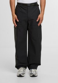 Schwarze, weit geschnittene Cargo-Hosen aus strapazierfähigem Stoff, mit mehreren Taschen und einem Gürtel. Getragen mit hellgrauen Sneakers.