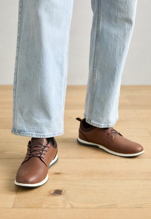 Chaussures en cuir marron avec des accents noirs, bouts arrondis et semelles en caoutchouc blanches, associées à un jean bleu clair à jambes larges sur un sol en bois.