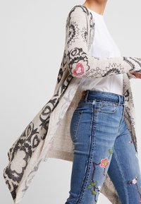 Lichte open voorkant cardigan in beige met zwarte bloemenpatronen en roze accenten. Gedragen over een wit shirt en gecombineerd met blauw geborduurde jeans.