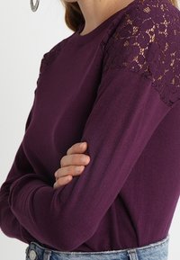 Kobieta w fioletowej bluzce z długim rękawem i koronkowymi detalami kwiatowymi na ramionach, z założonymi rękami, w połączeniu z jasnoniebieskimi dżinsami.