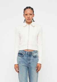 Pull en maille blanche cropped avec un col rond et une fermeture éclair à l'avant. Associé à un jean taille haute bleu clair, présentant un look décontracté.