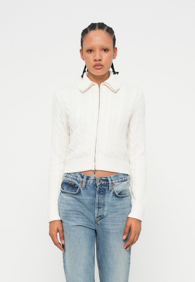 Pull en maille blanche cropped avec un col rond et une fermeture éclair à l'avant. Associé à un jean taille haute bleu clair, présentant un look décontracté.