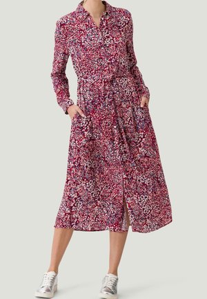 Vrouw die een lange mouw, gebloemde jurk met knopen draagt, voorzien van een taillekoord en zakken, gecombineerd met witte sneakers tegen een neutrale achtergrond.