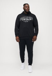 Schwarzer Hoodie mit Kapuze und weißem Textlogo, kombiniert mit passenden schwarzen Jogginghosen und weißen Sneakers. Einfaches Design, Material aus Baumwolle.