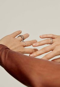 Dos manos adornadas con anillos se están acercando entre sí. Una mano tiene varios anillos de plata texturizados, mientras que la otra lleva una simple banda de plata.