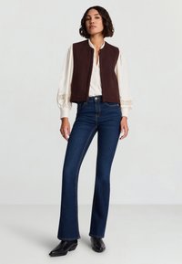 Braune ärmellose Weste über einer weißen Bluse mit Spitzenakzenten, kombiniert mit dunkelblauen Schlagjeans und schwarzen Stiefeln vor hellem Hintergrund.