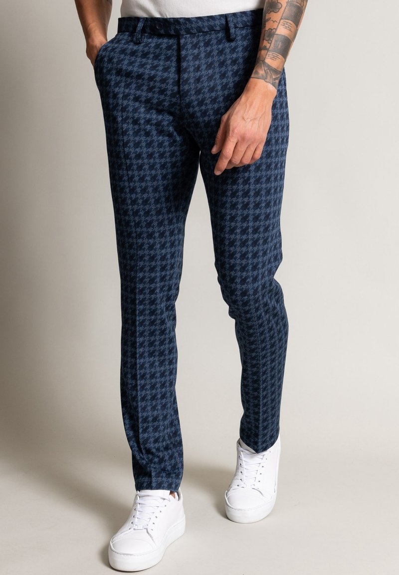 Homme portant un pantalon slim bleu marine à motif pied-de-poule et des baskets blanches, debout avec une main dans la poche, tatouage visible sur l'avant-bras.
