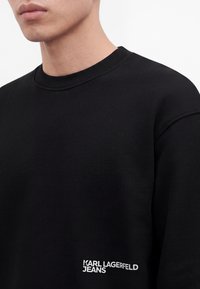 Близък план на човек, носещ черен суитчър с бял текст "Karl Lagerfeld Jeans" в долния десен преден сектор.