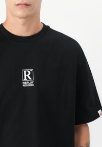 Black cotton t-shirt with short sleeves, featuring a white graphic logo of "R" and "REPLAY RECORDS" on the chest. No additional patterns or accents.