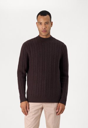 Pulover din tricot de culoare maro închis, cu un guler înalt cu nervuri, având modele texturate și manșete și tiv cu nervuri. Asortat cu pantaloni bej deschis.