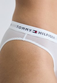 Fehér csíkos alsónemű, amelyen a "TOMMY HILFIGER" logós szalag található. Az anyag simának tűnik, puha tapintású és finom fénylik.