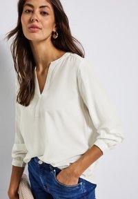 Femme aux longs cheveux bruns portant des boucles d'oreilles en or, un chemisier blanc à manches longues, un jeans bleu avec une main dans la poche, tenant un sac tissé.