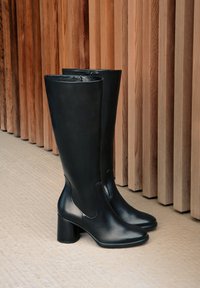 Pair of black leather knee-high boots with block heels placed on textured floor against wooden vertical slat wall.