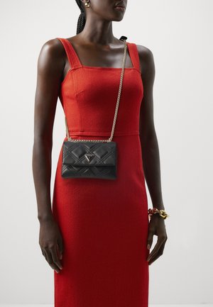 Femme portant une robe rouge sans manches avec un sac bandoulière noir matelassé et un gros bracelet doré et rouge à son poignet droit.
