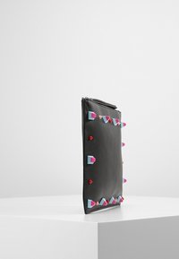 Czarna prostokątna torebka kopertowa z kolorowymi geometrycznymi ćwiekami w kolorach niebieskim, różowym i czerwonym, na białej powierzchni na tle jednolitej białej ściany.