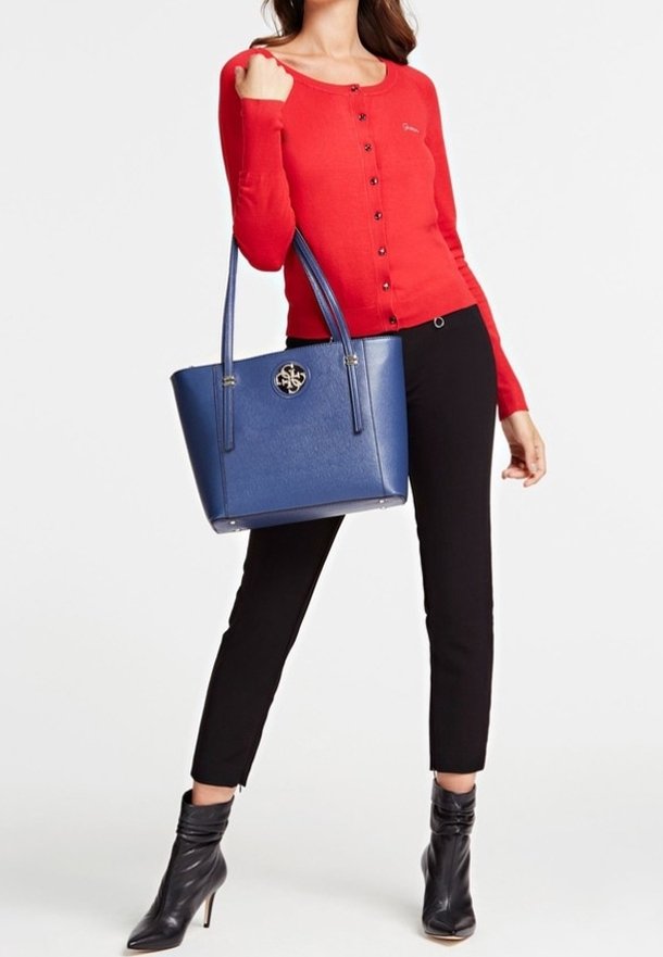 Femme portant un haut rouge boutonné, un pantalon noir et des bottines noires, portant un grand sac cabas bleu avec un logo rond argenté.