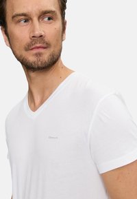 Weißes V-Ausschnitt-T-Shirt aus weichem Stoff, mit kurzen Ärmeln und einem kleinen grauen Logo auf der Brust. Einfaches, leichtes Design.