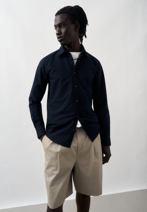 Άνδρας με σκούρα πλεγμένα μαλλιά φοράει σκούρο πουκάμισο με κουμπιά πάνω από λευκή μπλούζα και μπεζ σορτς μέχρι το γόνατο, στέκεται μπροστά σε απλό ανοιχτό φόντο.