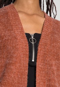 Cardigan lavorato a maglia ruggine con una texture a coste, caratterizzato da una ampia scollatura a V e apertura frontale, indossato sopra un top nero con zip.