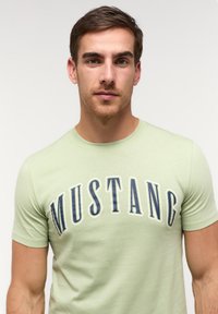 Világoszöld póló, amelyen a "MUSTANG" felirat zsírkrétaszerű, sötétkék betűkkel szerepel. Az anyag lágy és sima tapintású, rövid ujjú.