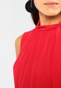 Czerwony top bez rękawów z plisowanym wzorem i wysokim kołnierzem. Modelka nosi duże okrągłe czarne kolczyki ze złotym akcentem.