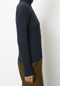 Langärmliges Rollkragenoberteil in tiefem Marineblau, aus dehnbarem Stoff. Figurbetonter Schnitt mit glatter Textur und minimalen Nähten. Kombiniert mit olivgrünen Hosen.