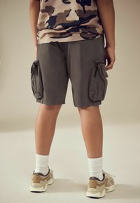 Shorts cargo gris avec poches latérales, associés à des sneakers beiges et des chaussettes blanches. La chemise arbore un imprimé camouflage beige et marine.