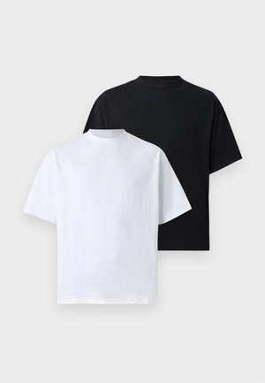 Due giovani adulti in piedi, indossano magliette nere larghe coordinate, pantaloni neri e scarpe nere robuste, su uno sfondo bianco semplice.