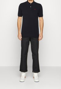 Polo noir avec trois boutons, logo brodé sur la poitrine gauche, associé à un pantalon noir et des baskets blanches avec des accents.