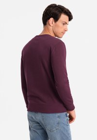 Sweatshirt bordeaux à manches longues avec un col rond et des poignets côtelés. Porté par-dessus un jean bleu clair, montrant une texture douce en coton.