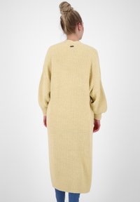 Długi, dziergany kardigan w miękkim żółtym kolorze z prążkowaną fakturą, rękawami z opuszczonymi ramionami oraz otwarciem na plecach w pobliżu dekoltu.
