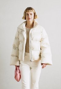 Krémová nafukovací bunda, která má zipový uzávěr, vysoký límec a velké prošívané části. V kombinaci se světlými kalhotami a růžovou kabelkou.