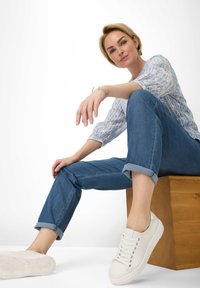 White sneakers with laces, blue rolled-up jeans, and a light patterned blouse, seated on a wooden block against a plain backdrop.